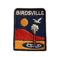 Birdsville embroidered travel patch featuring artesian bore releasing water against orange desert dunes, a golden sun and a flying bird.
