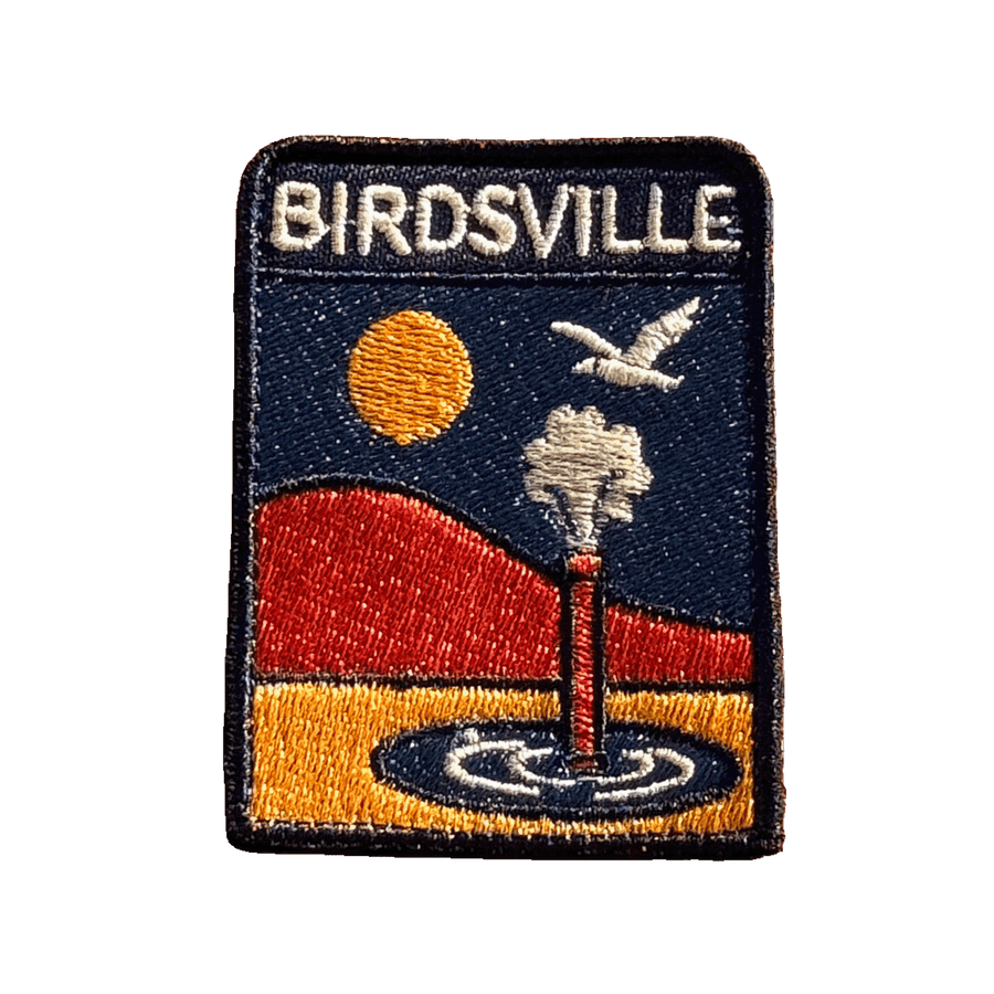 Birdsville embroidered travel patch featuring artesian bore releasing water against orange desert dunes, a golden sun and a flying bird.