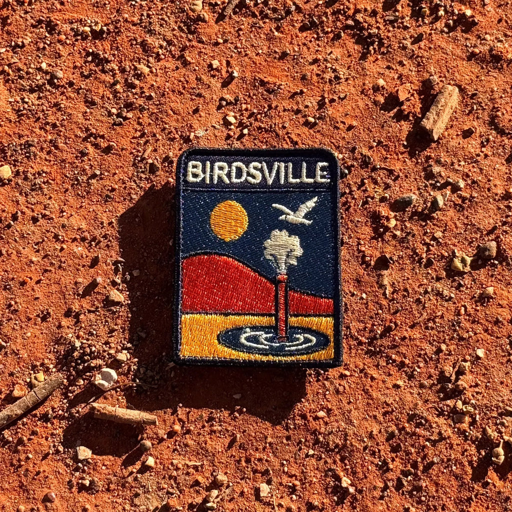 Birdsville embroidered travel patch featuring artesian bore releasing water against orange desert dunes, a golden sun and a flying bird.