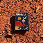 Birdsville embroidered travel patch featuring artesian bore releasing water against orange desert dunes, a golden sun and a flying bird.