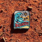 Bondi Beach embroidered travel patch featuring four surfers riding turquoise ocean waves beside a sunny golden beach.