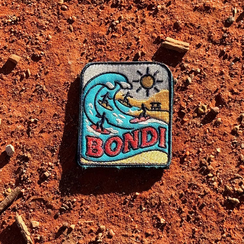 Bondi Beach embroidered travel patch featuring four surfers riding turquoise ocean waves beside a sunny golden beach.