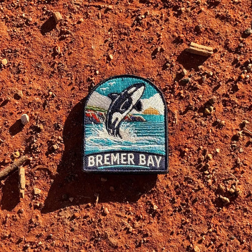 Bremer Bay embroidered travel patch featuring breaching orca splashing in the ocean near a rocky coastline at sunset.