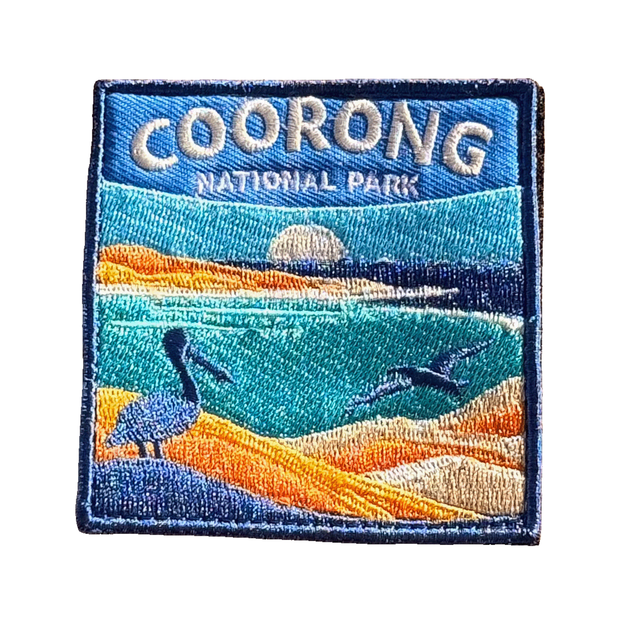 Coorong National Park embroidered travel patch with a pelican on sand dunes and a dolphin in turquoise coastal water; photographed on a transparent background.