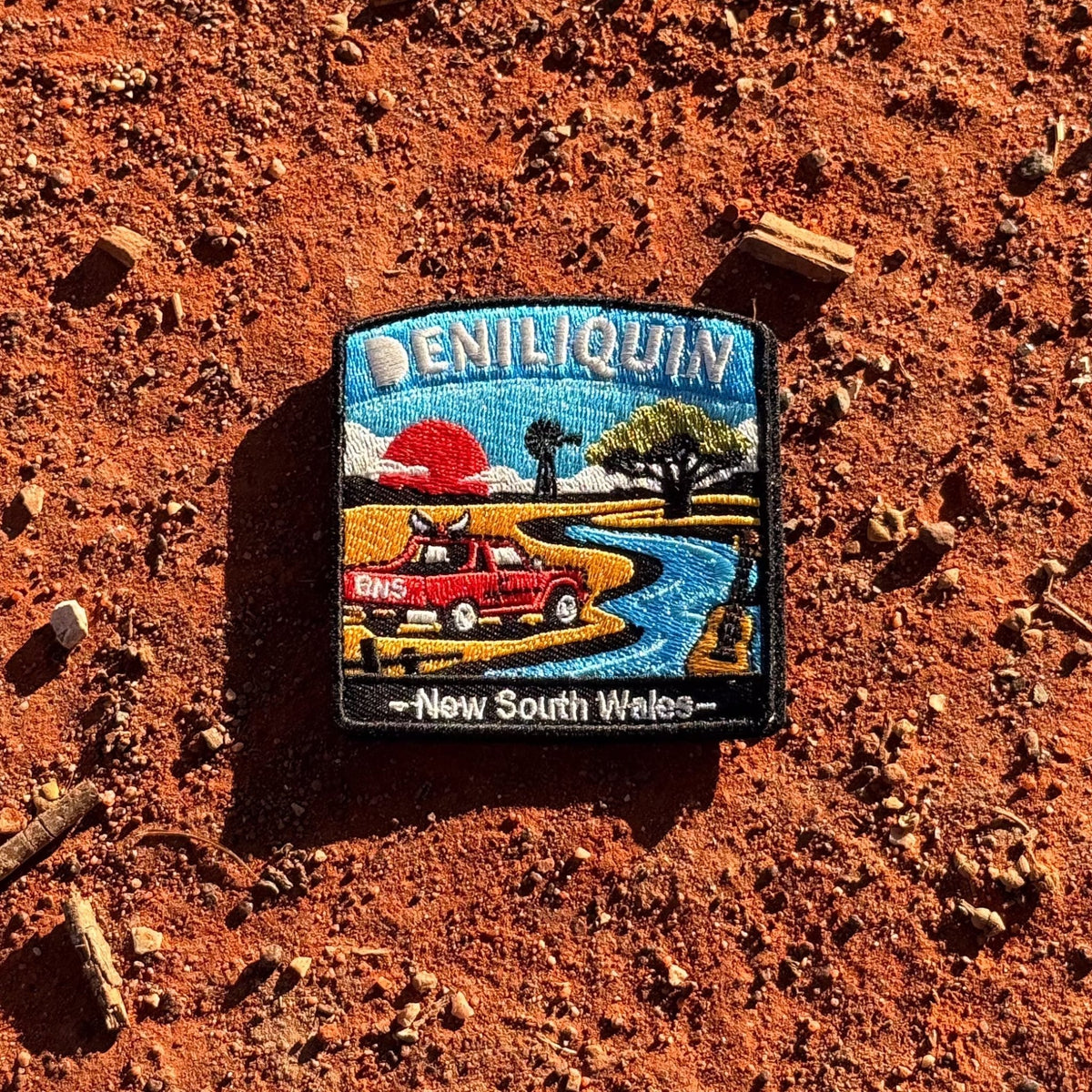 Deniliquin embroidered travel patch with a red ute, guitar, windmill and winding river beneath an orange sunset.