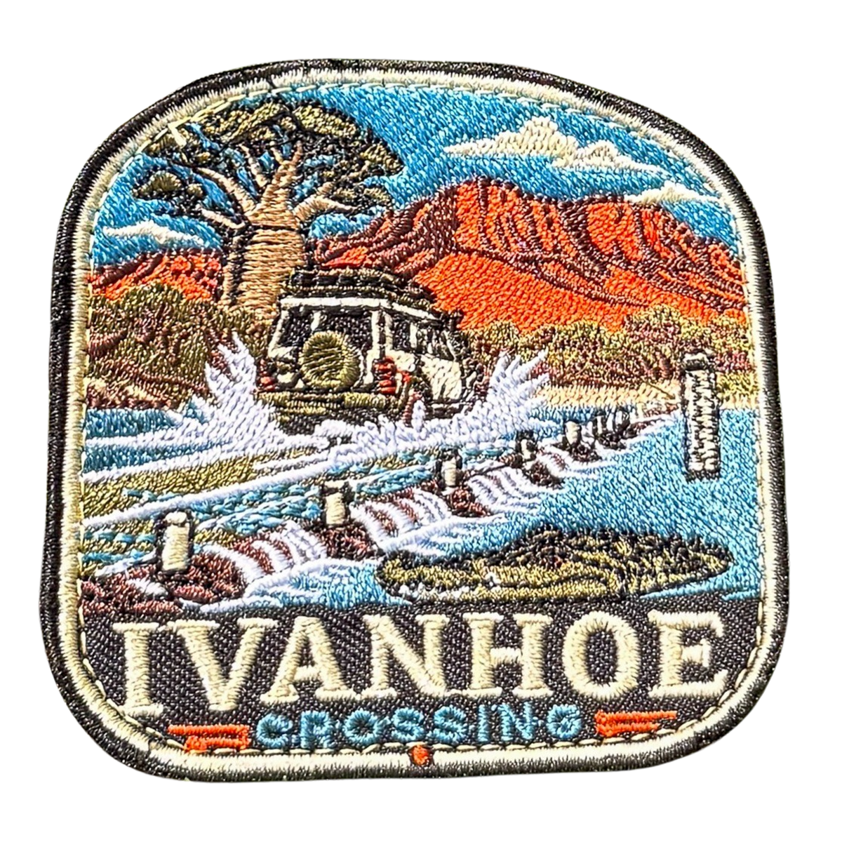 Ivanhoe Crossing embroidered travel patch with a four-wheel drive splashing across a flooded causeway with a crocodile in the water; photographed on a transparent background.