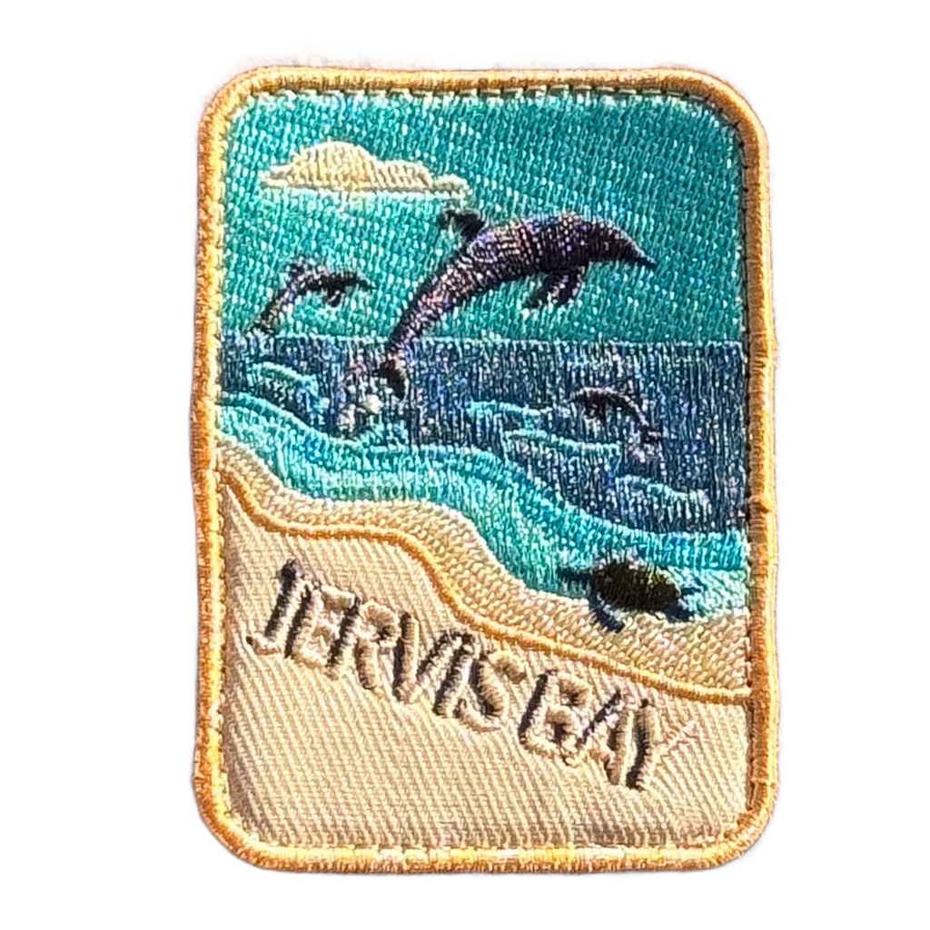 Jervis Bay embroidered travel patch with dolphins leaping offshore and a turtle swimming beside a sandy beach; photographed on a transparent background.