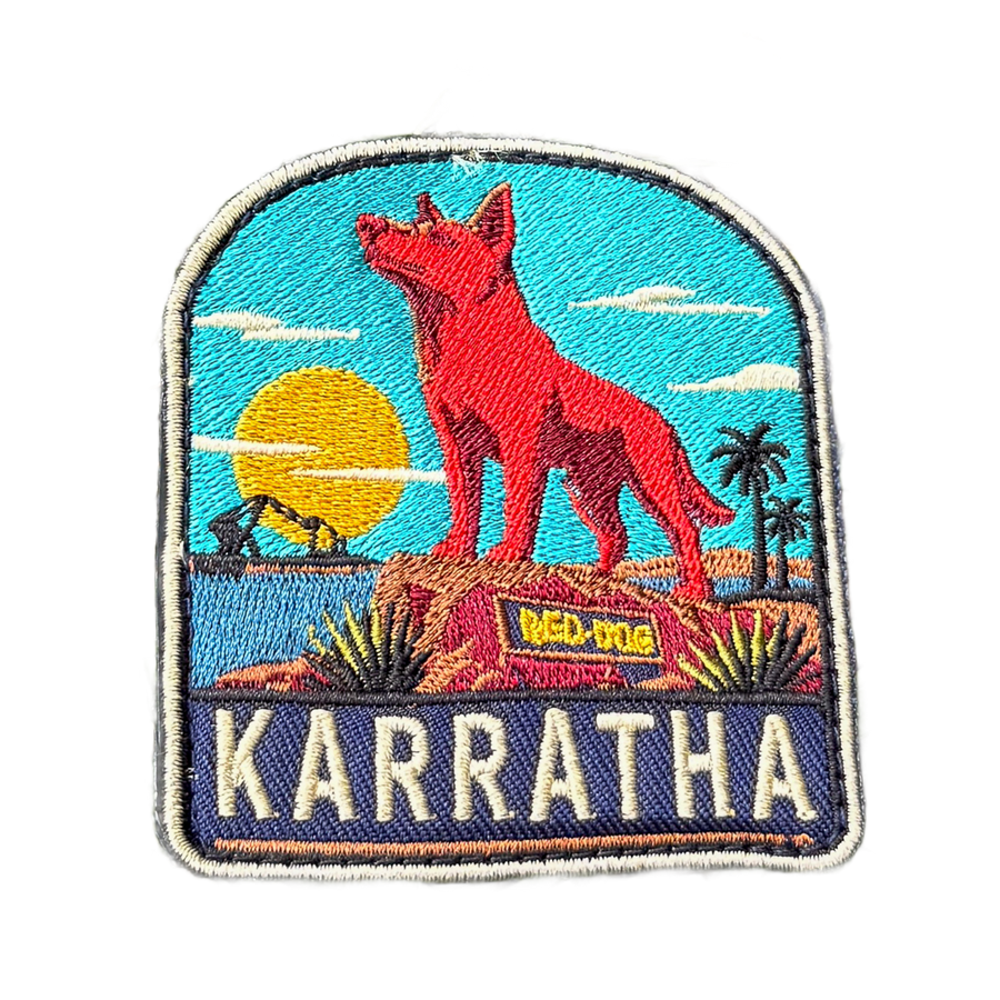 Karratha embroidered travel patch with the Red Dog statue on a red rocky hill with an industrial harbour and sunset; photographed on a transparent background.