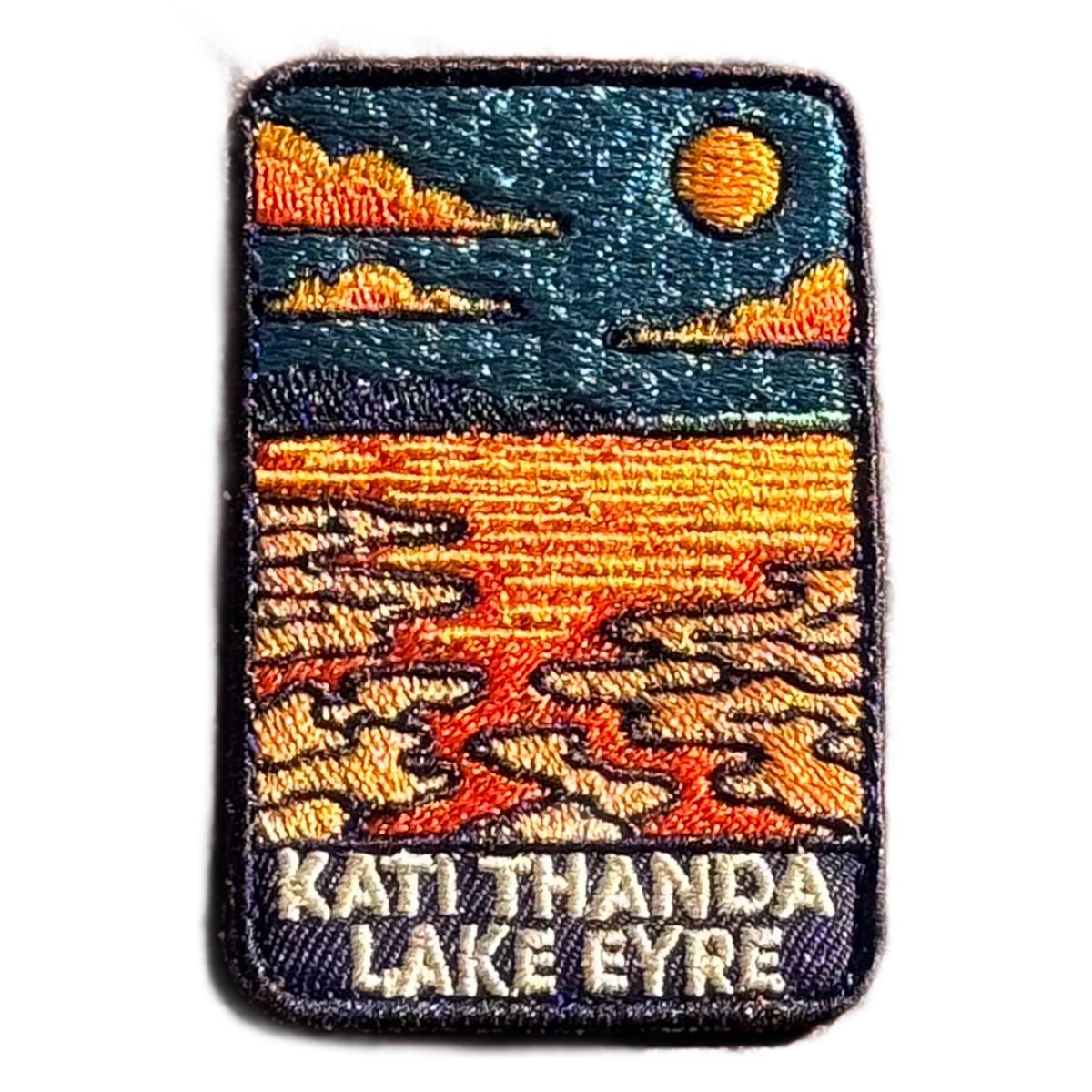 Lake Eyre (Kati Thanda) embroidered travel patch with cracked salt flats and shallow water beneath an orange sun at Kati Thanda‚ÄìLake Eyre; photographed on a transparent background.