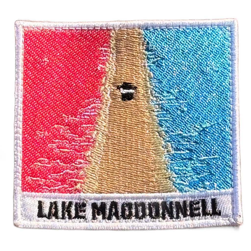 Lake MacDonnell embroidered travel patch with an aerial view of a road separating bright pink and blue lakes; photographed on a transparent background.