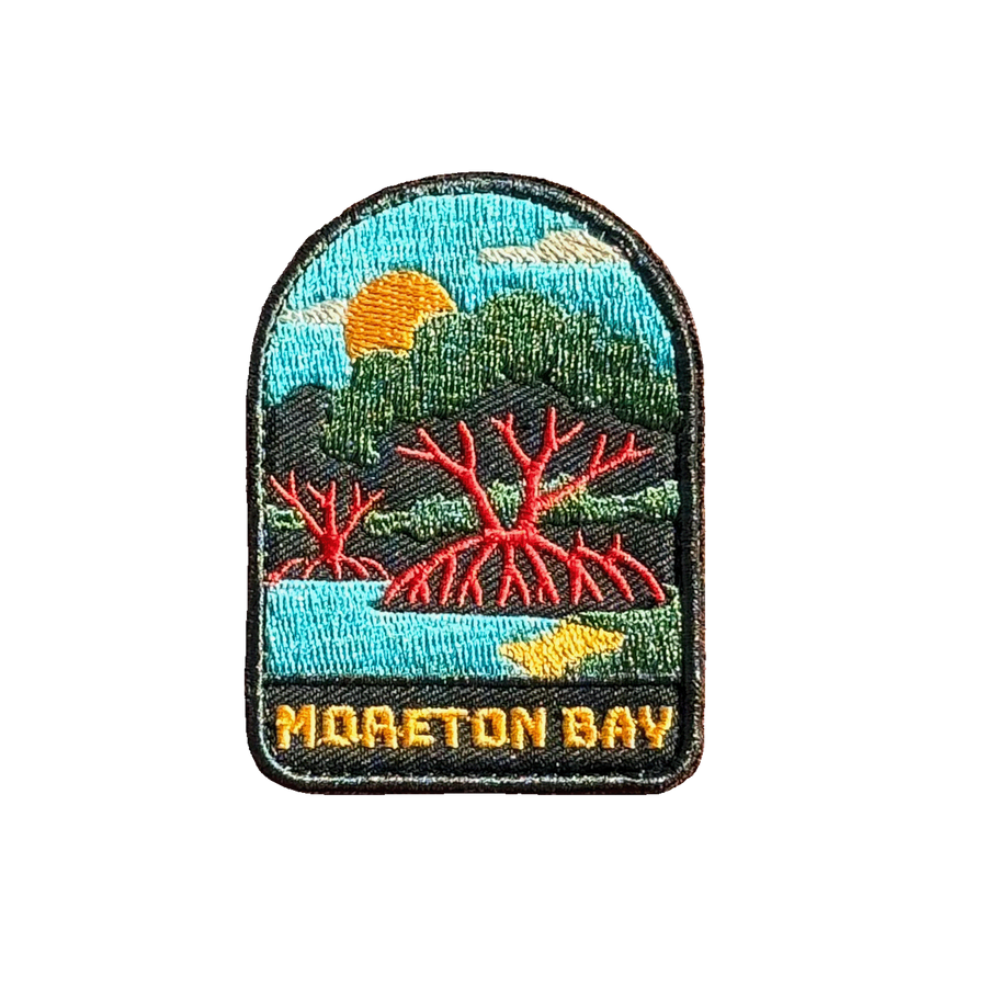 Moreton Bay embroidered travel patch with mangrove trees with exposed roots beside calm coastal water at sunset; photographed on a transparent background.