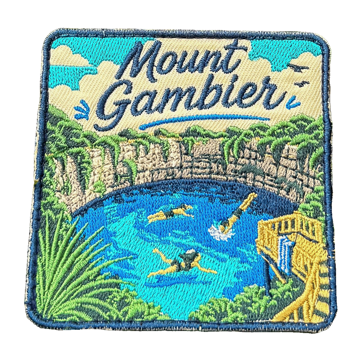 Mount Gambier embroidered travel patch with swimmers in a brilliant blue crater lake surrounded by limestone cliffs; photographed on a transparent background.