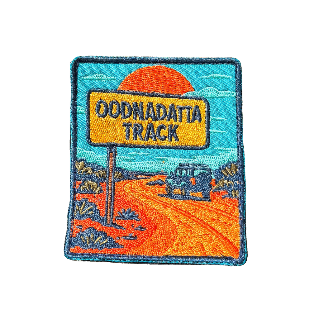 Oodnadatta Track embroidered travel patch with an Oodnadatta Track roadside sign and four-wheel drive on a red dirt road at sunset; photographed on a transparent background.