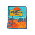 Oodnadatta Track embroidered travel patch with an Oodnadatta Track roadside sign and four-wheel drive on a red dirt road at sunset; photographed on a transparent background.