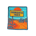 Oodnadatta Track embroidered travel patch with an Oodnadatta Track roadside sign and four-wheel drive on a red dirt road at sunset; photographed on a transparent background.