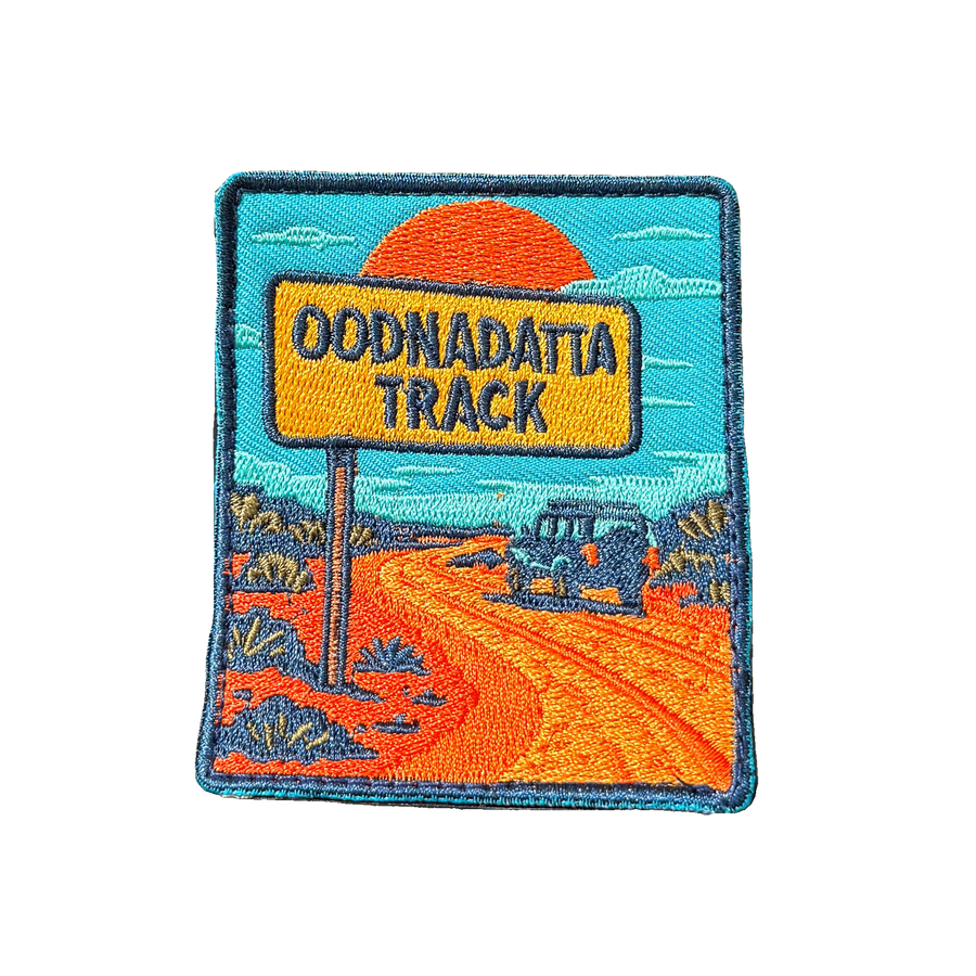 Oodnadatta Track embroidered travel patch with an Oodnadatta Track roadside sign and four-wheel drive on a red dirt road at sunset; photographed on a transparent background.