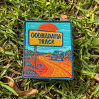Oodnadatta Track embroidered travel patch with an Oodnadatta Track roadside sign and four-wheel drive on a red dirt road at sunset.