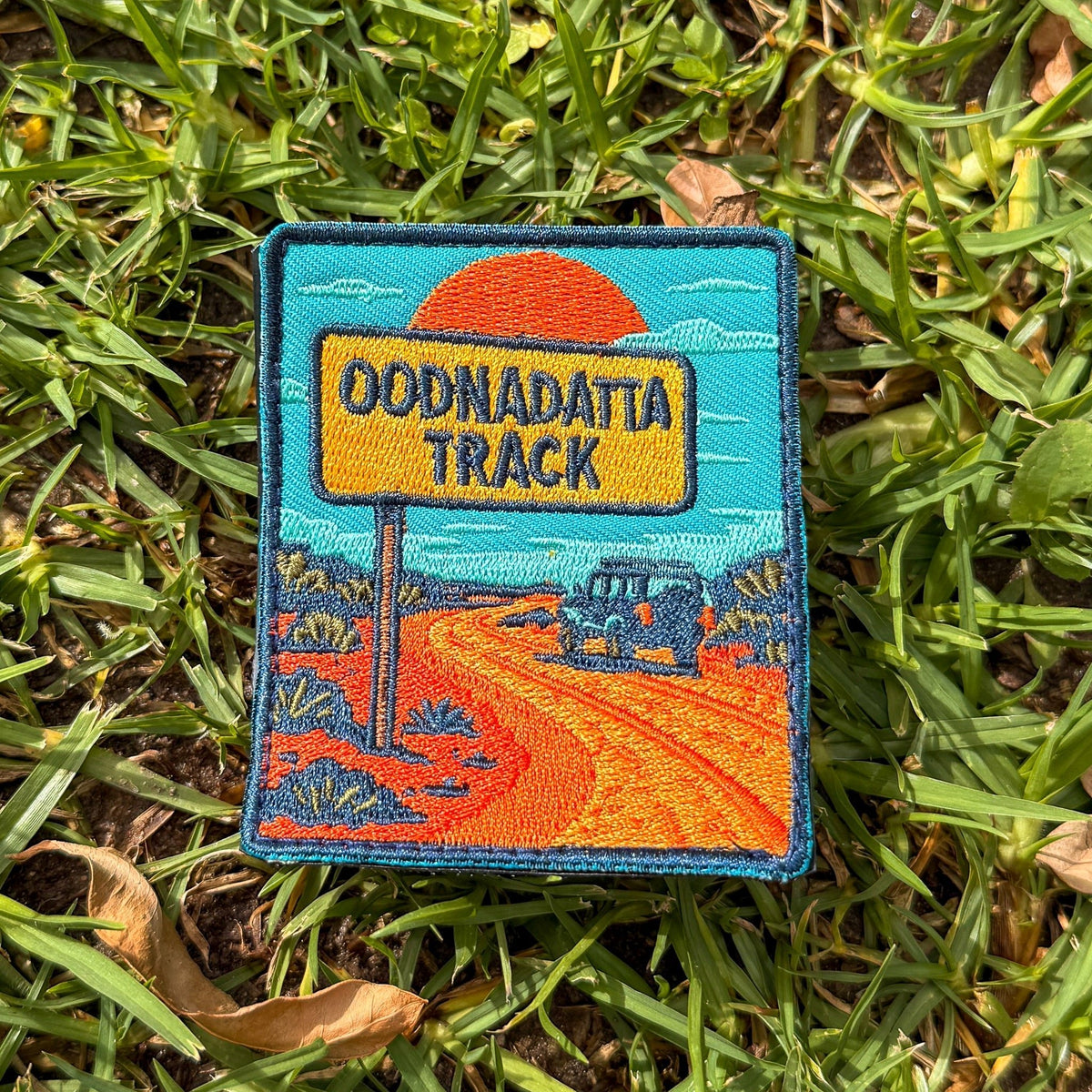 Oodnadatta Track embroidered travel patch with an Oodnadatta Track roadside sign and four-wheel drive on a red dirt road at sunset.