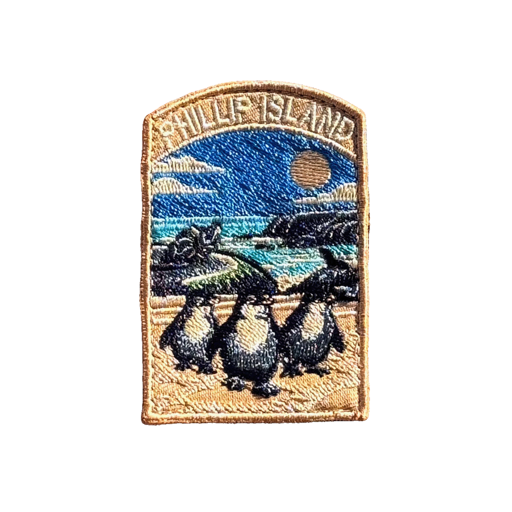 Phillip Island embroidered travel patch with three little penguins on a beach with a motorcycle racer and ocean cliffs behind; photographed on a transparent background.