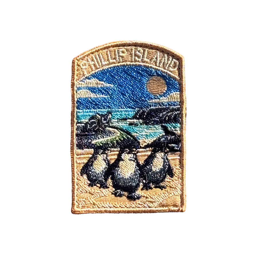 Phillip Island embroidered travel patch with three little penguins on a beach with a motorcycle racer and ocean cliffs behind; photographed on a transparent background.