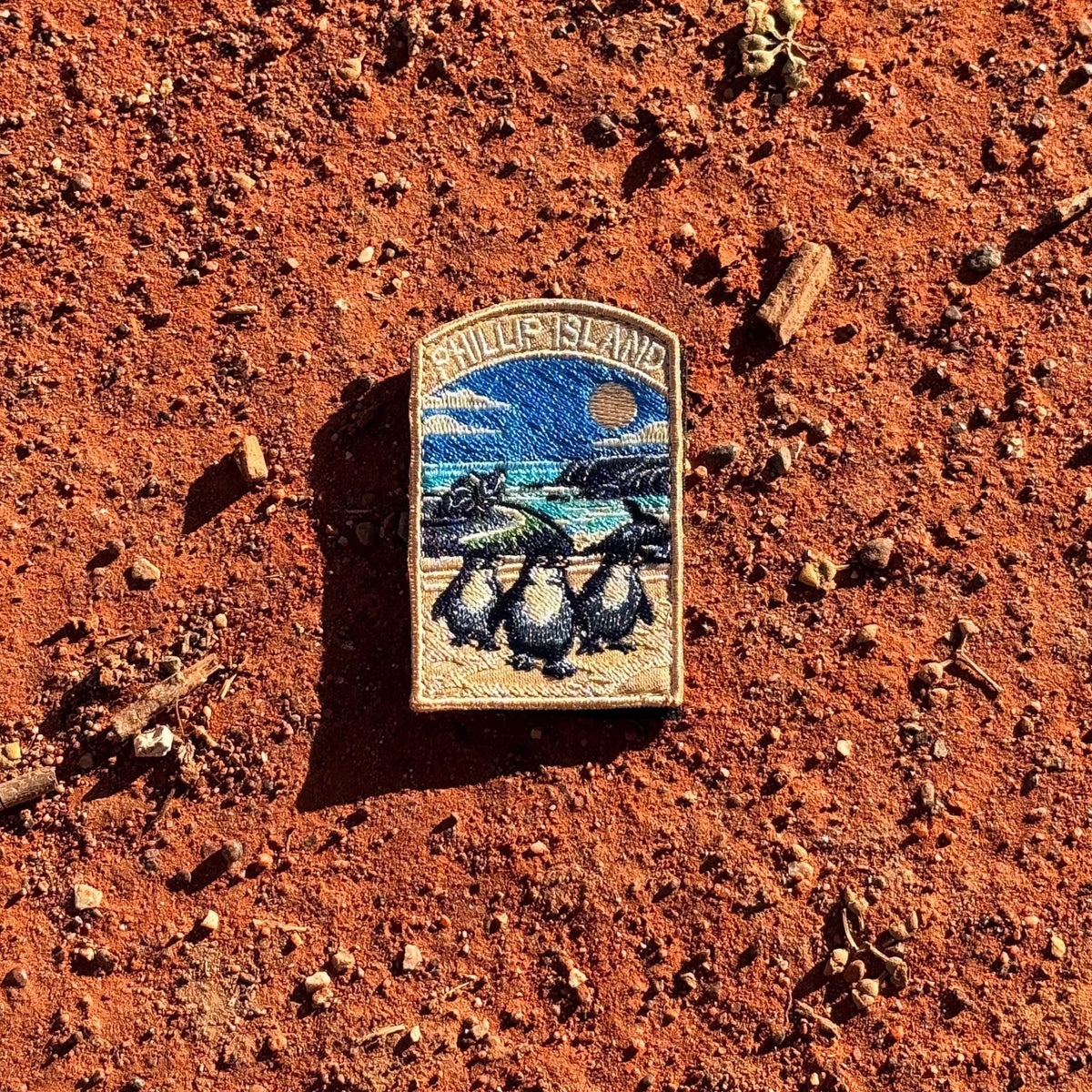 Phillip Island embroidered travel patch with three little penguins on a beach with a motorcycle racer and ocean cliffs behind.