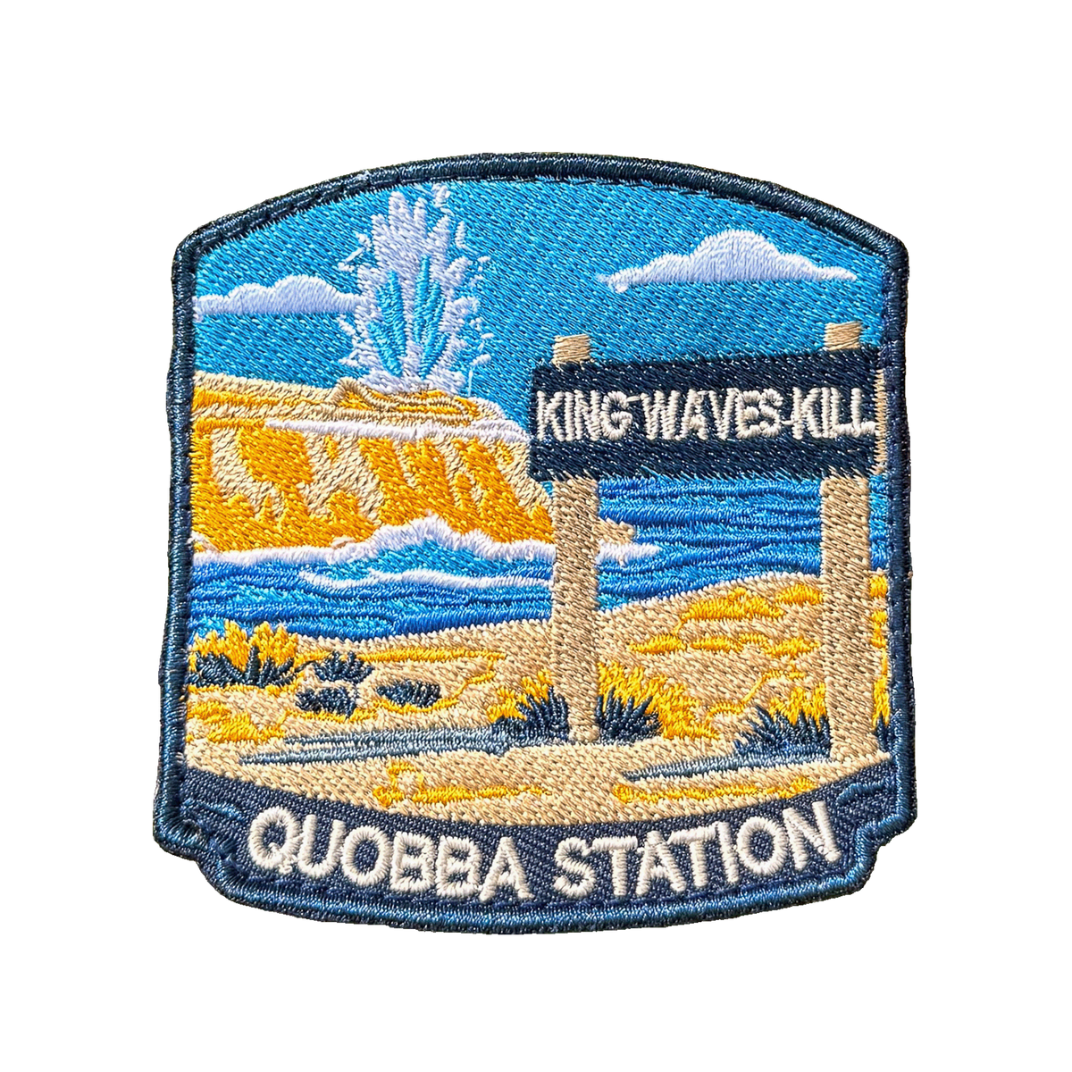 Quobba Blowholes embroidered travel patch with a coastal blowhole spraying above rugged cliffs and ocean; photographed on a transparent background.