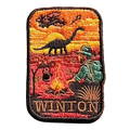 Winton embroidered travel patch with a dinosaur, vintage aircraft and traveller sitting beside a campfire at sunset; photographed on a transparent background.
