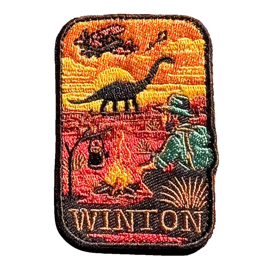 Winton embroidered travel patch with a dinosaur, vintage aircraft and traveller sitting beside a campfire at sunset; photographed on a transparent background.