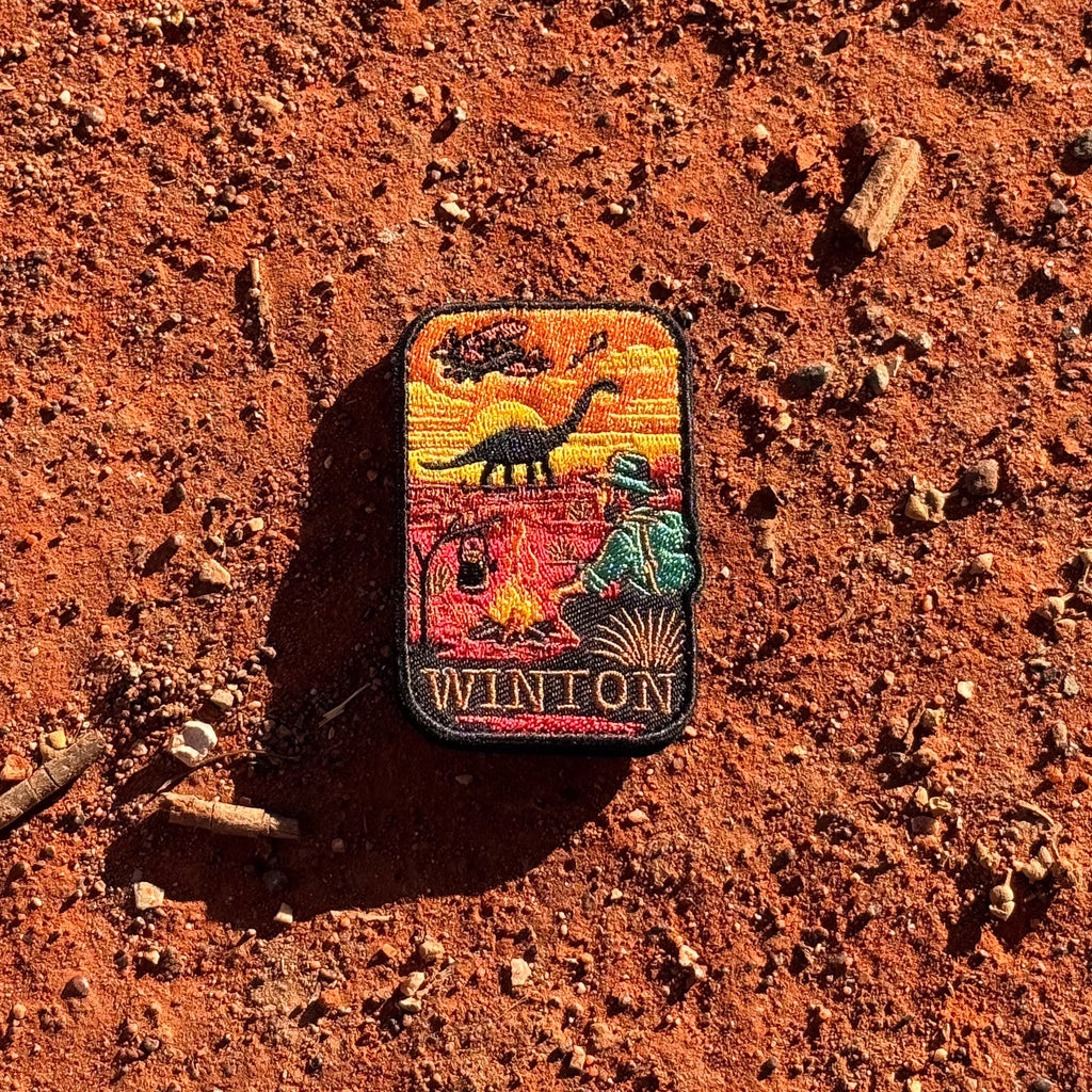 Winton embroidered travel patch with a dinosaur, vintage aircraft and traveller sitting beside a campfire at sunset.