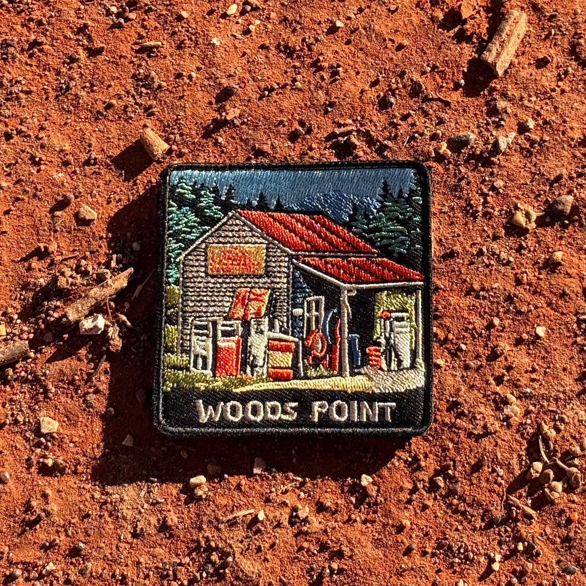 Woods Point embroidered travel patch with a rustic high-country petrol station with old fuel pumps among pine trees.
