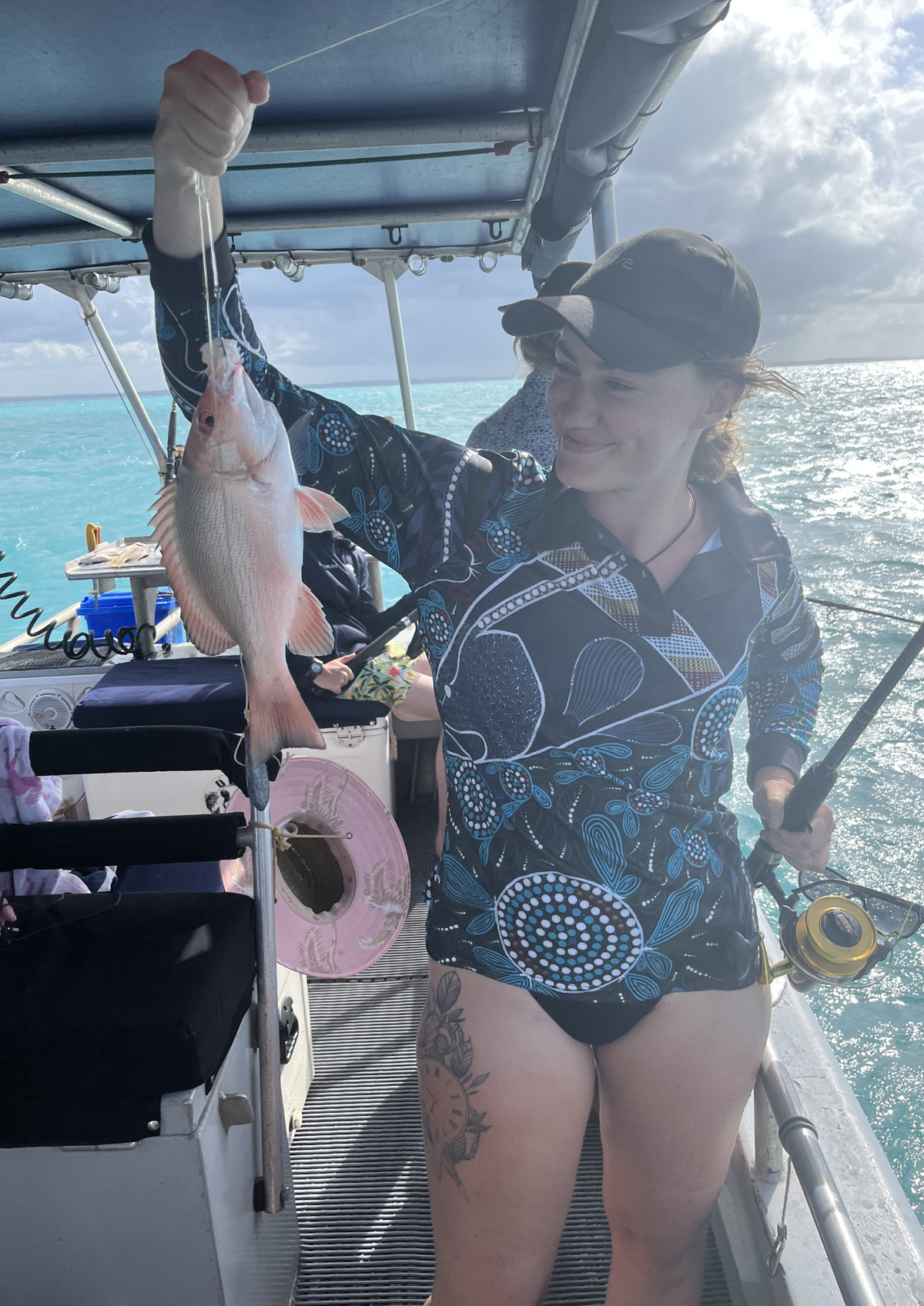 A woman on a boat holds up a freshly caught fish while wearing a dark patterned long-sleeve swimsuit and a black cap, with ocean water and cloudy skies in the background.