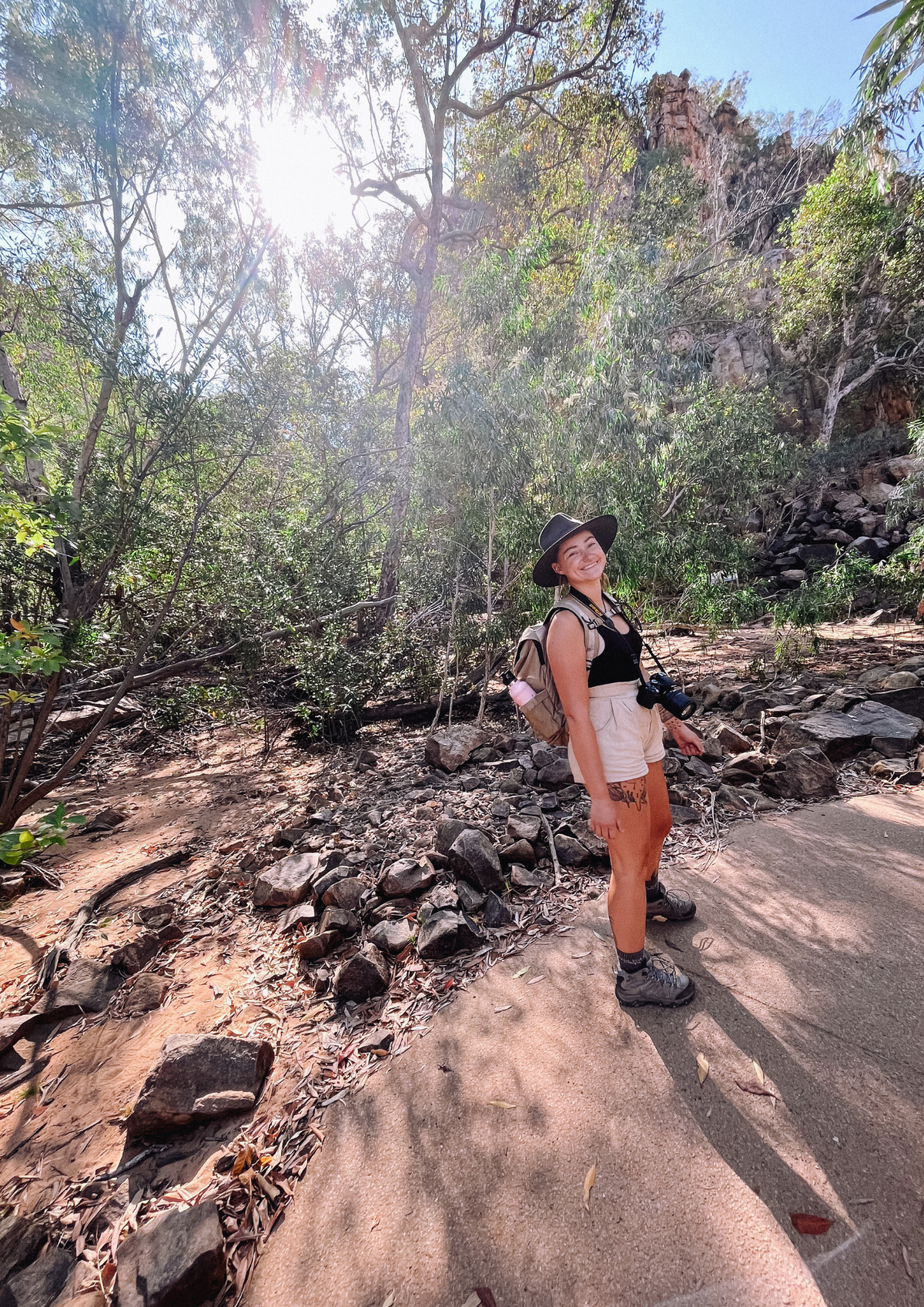 A woman wearing a black wide-brim Akubra hat, black sleeveless top, and cream shorts stands on a dirt and concrete trail in a sunlit forest clearing with rocks and greenery around her. This is a part of the Katherine Gorge Tour.