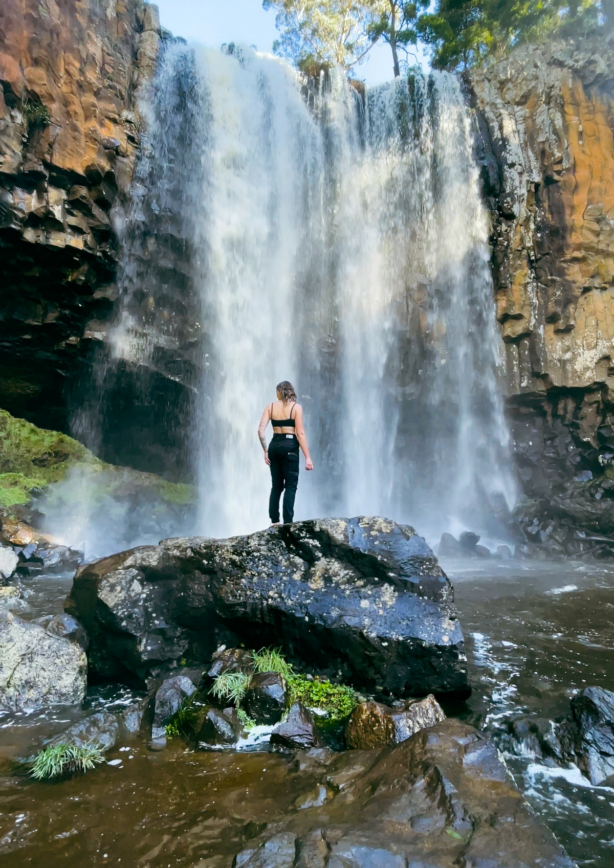A woman stands on a large rock in front of a tall waterfall in a rocky natural setting, wearing a black sports bra and black FXD pants..