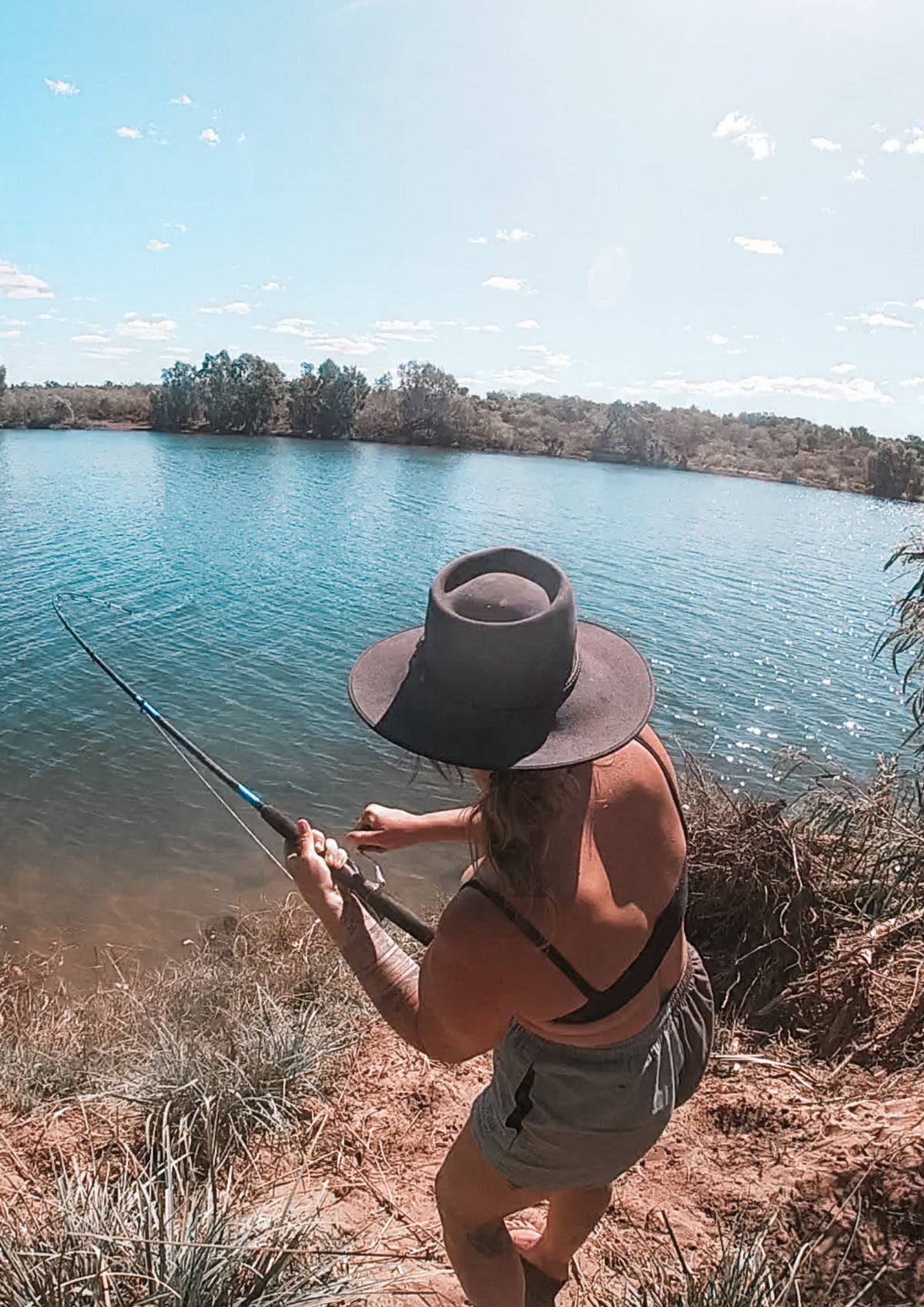 A woman wearing a wide-brimmed dark hat, a black strappy top, and gray shorts stands on a riverbank holding a fishing rod, with a river and trees in the background. This is Seven Emu Station.