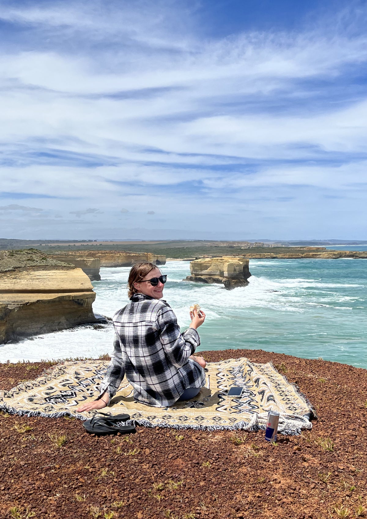 A woman sits cross-legged on a patterned blanket on a rocky cliff overlooking the ocean, wearing a black and white plaid flannel shirt and sunglasses, with a scenic coastal landscape featuring limestone stacks and turquoise water under a blue sky.