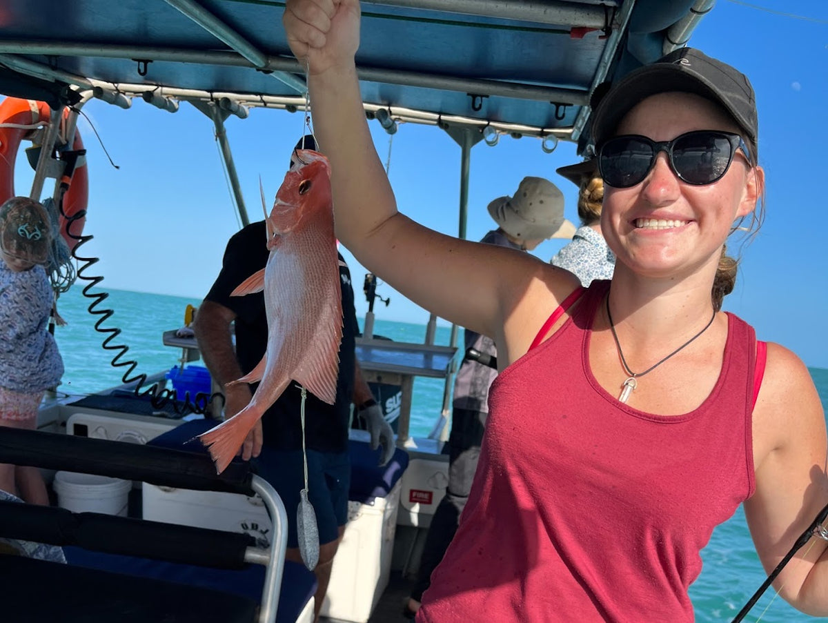 A woman on a boat wearing a red tank top and sunglasses holds up a pinkish-red fish she caught, with ocean water and other people visible in the background.