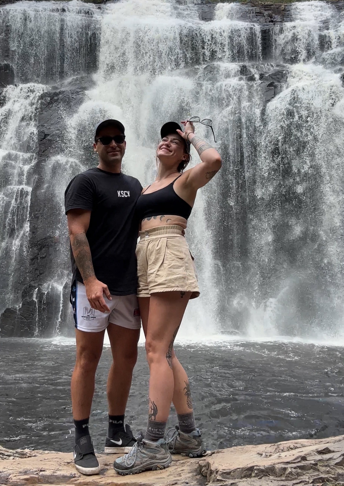 A man and a woman posing together in front of a large waterfall, with the man wearing a black t-shirt and white shorts and the woman wearing a black crop top and beige shorts.