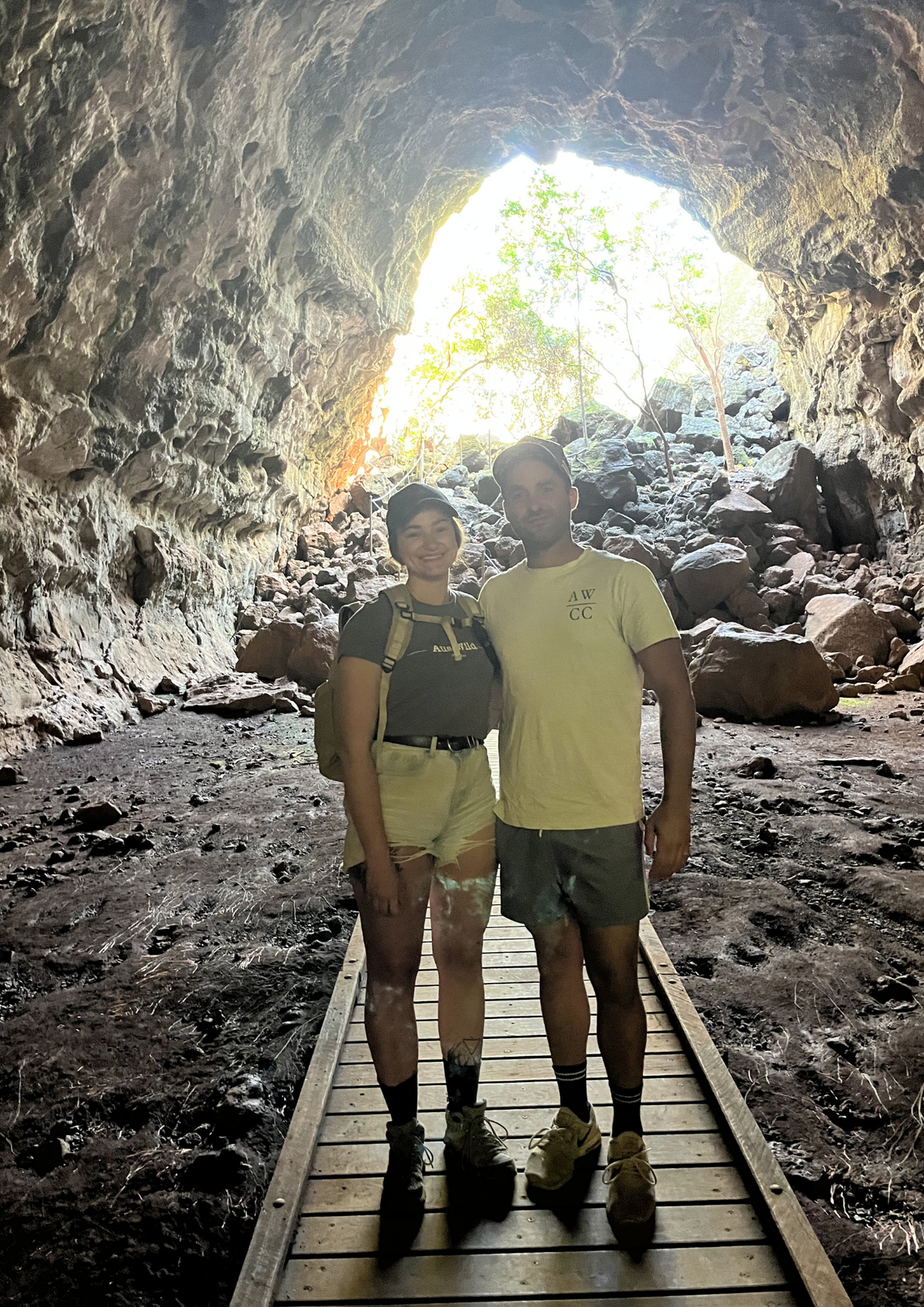 Two people standing on a wooden boardwalk inside a rocky cave, with a bright opening of daylight and greenery visible in the background. The Cave is a part of the Undara Lava Tubes Tour in Outback Queensland.