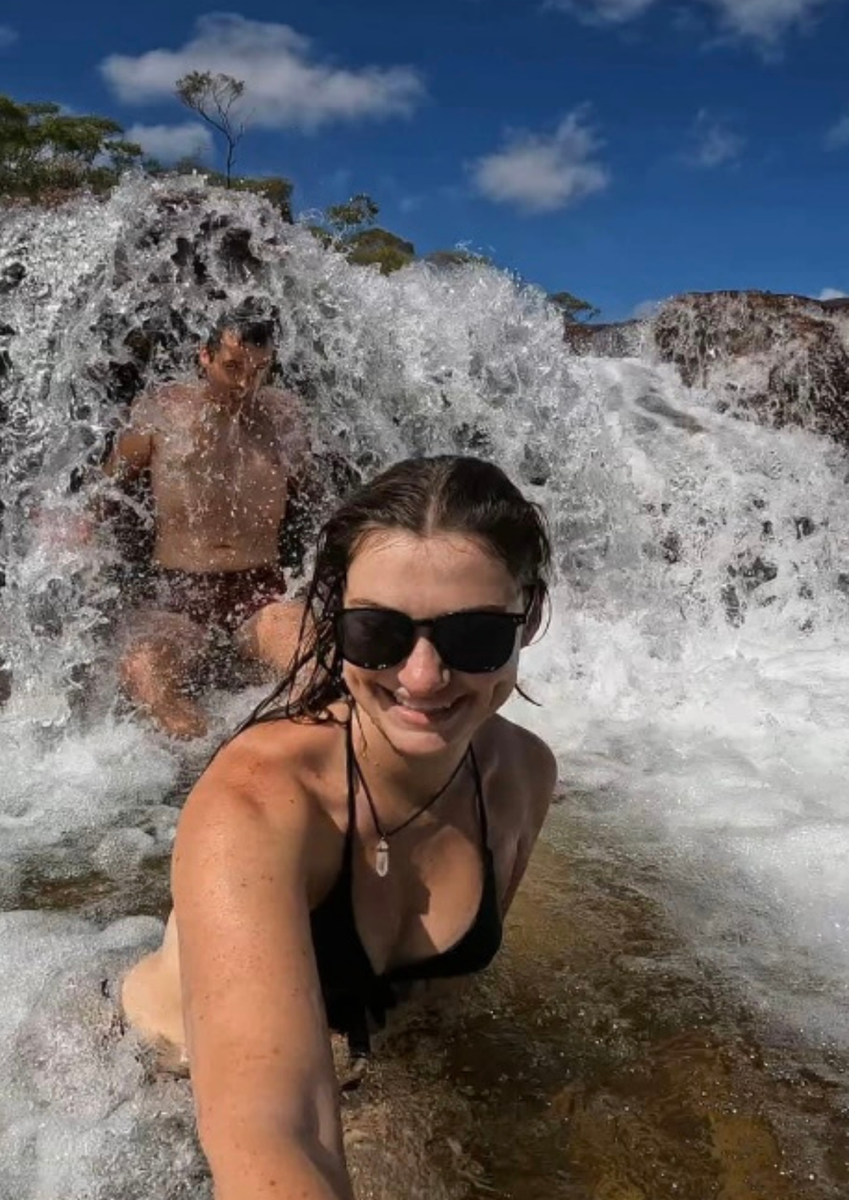 A woman in a black bikini and sunglasses takes a selfie while sitting in the rushing water of a small waterfall, with a man sitting behind her under the cascading water.