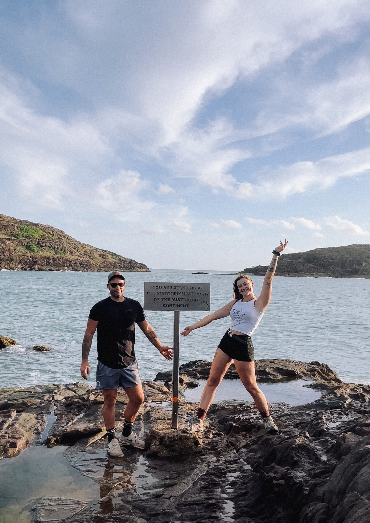 A man and a woman posing on dark rocks by the ocean, with a signpost between them and a rocky coastline under a cloudy blue sky in the background. This is the Northern Most Point of Mainland Australia - Cape York.