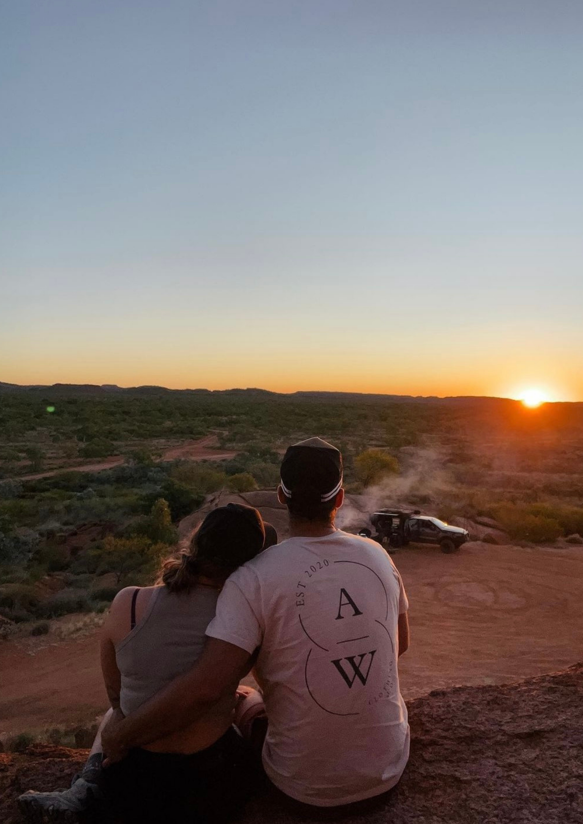 A couple sits on a rocky cliff overlooking a desert landscape at sunset, with a vehicle parked in the distance. The man wears a white graphic t-shirt with a black circular logo featuring the letters 'A' and 'W'.