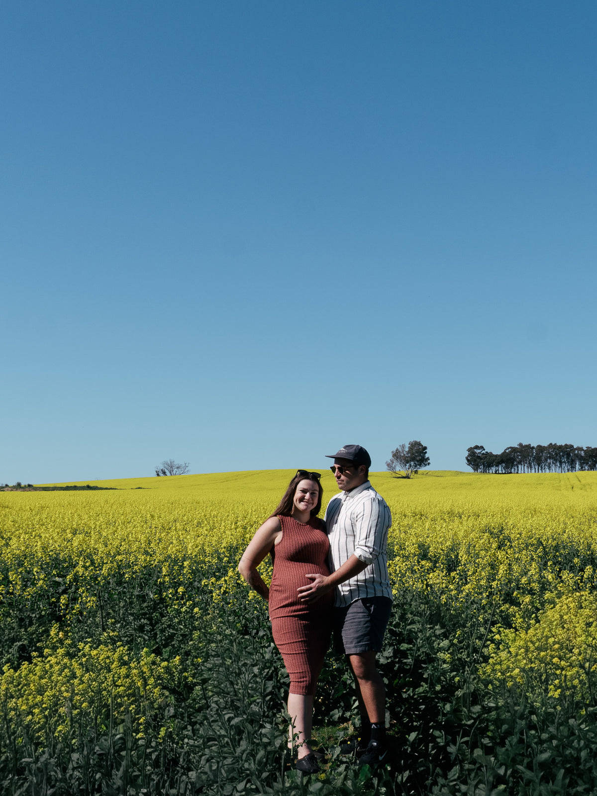A couple posing in a bright yellow flower (Canola) field under a clear blue sky. The woman is pregnant, wearing a sleeveless rust-brown dress, and the man is wearing a white shirt, dark shorts, and a cap.