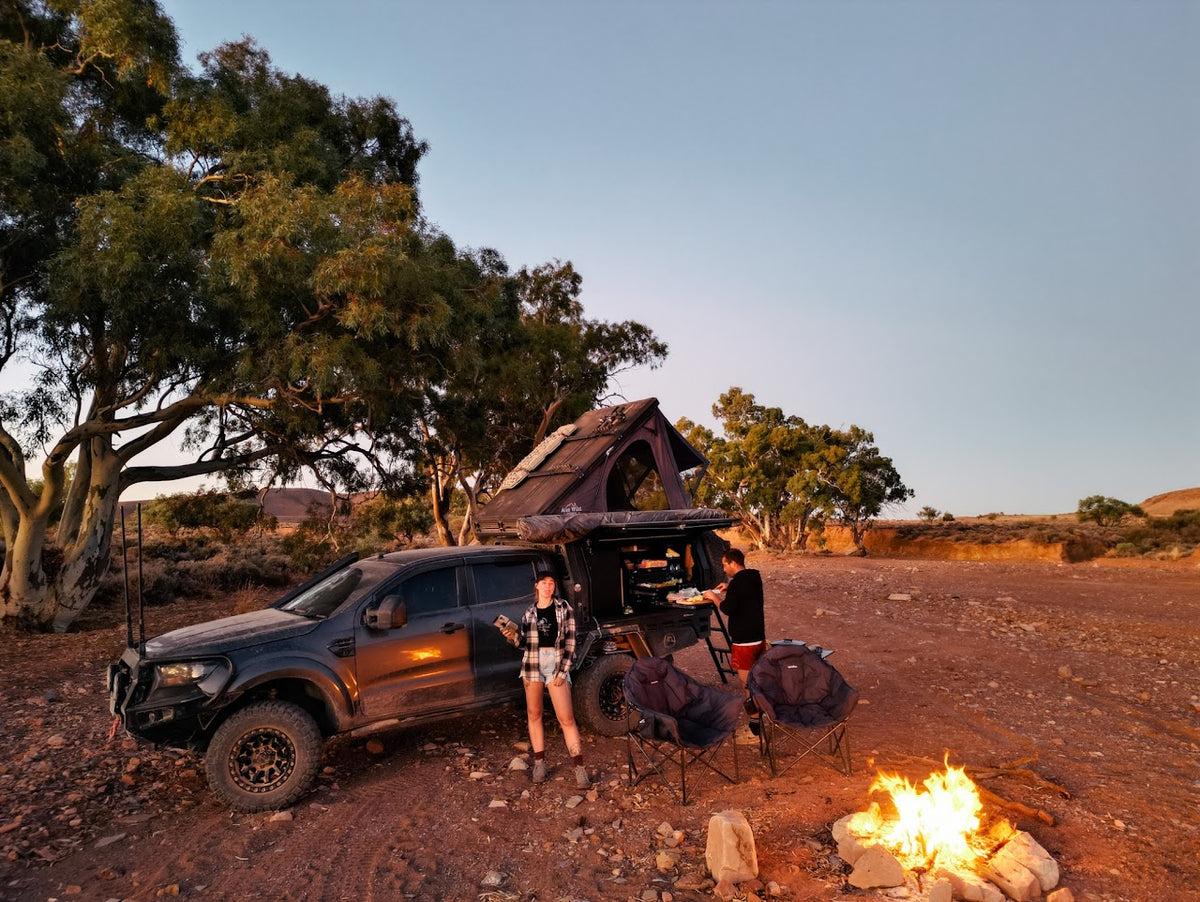 A black off-road 4wd with a rooftop tent is parked on a dirt clearing at dusk, with two people standing beside it next to camping chairs and a small campfire. Trees and open land are visible in the background.