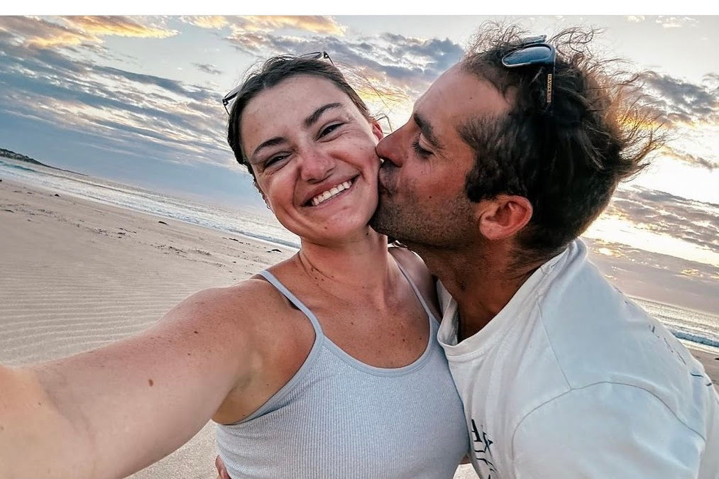 A smiling woman in a light gray ribbed tank top takes a selfie on a sandy beach while a man in a white t-shirt kisses her cheek, with a sunset sky and ocean in the background.