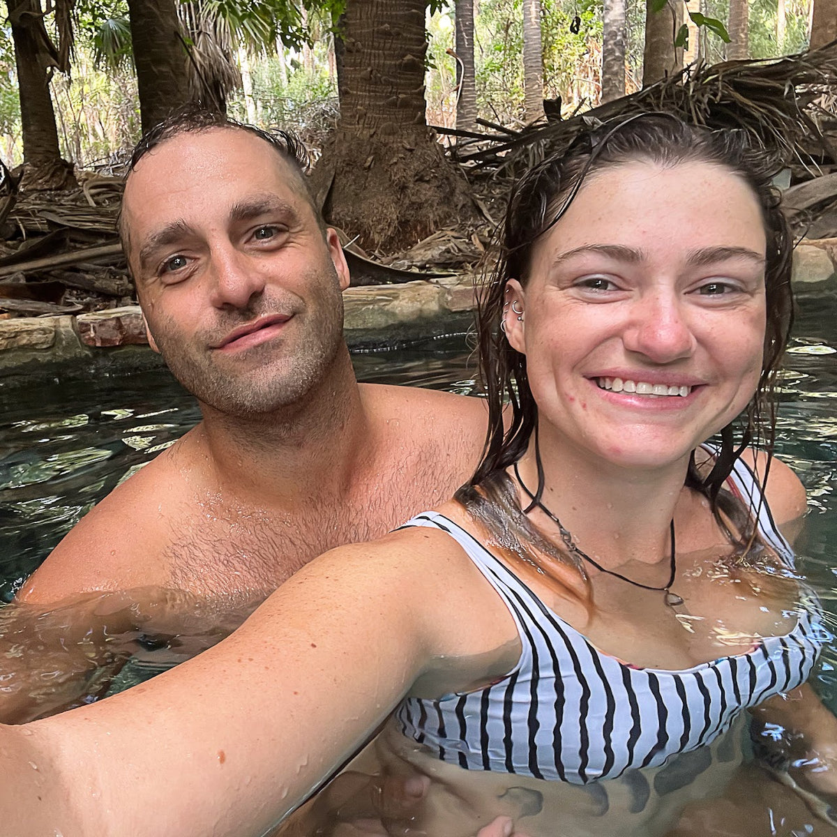 A smiling couple taking a selfie in a natural hot spring at Mataranka surrounded by trees and rocks. The woman wears a black and white striped bikini top, and the man is shirtless.