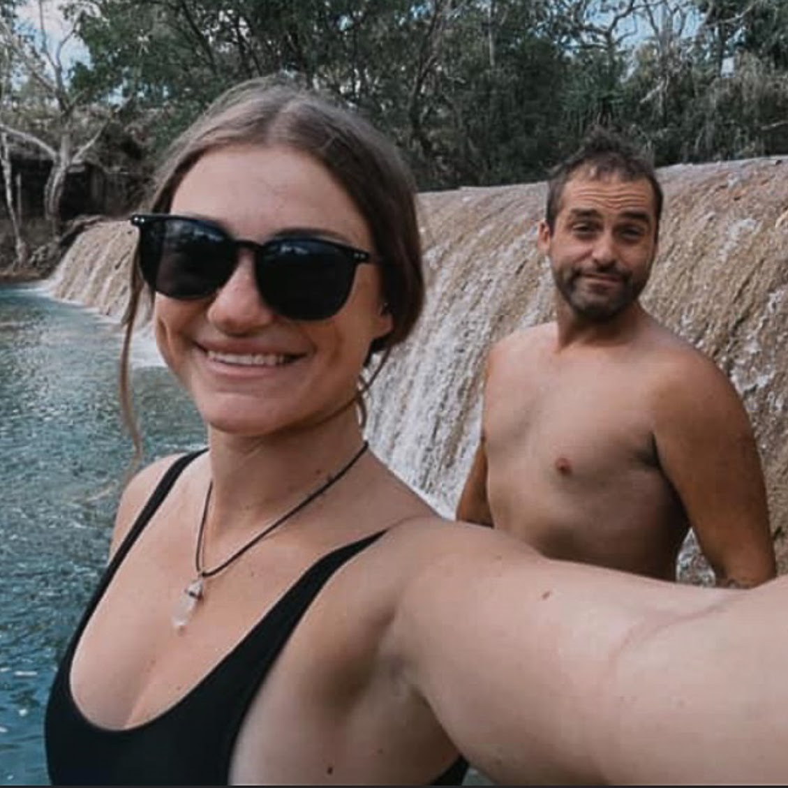 A smiling woman in a black swimsuit and sunglasses takes a selfie in front of a waterfall, with a shirtless man standing behind her in an outdoor natural setting.