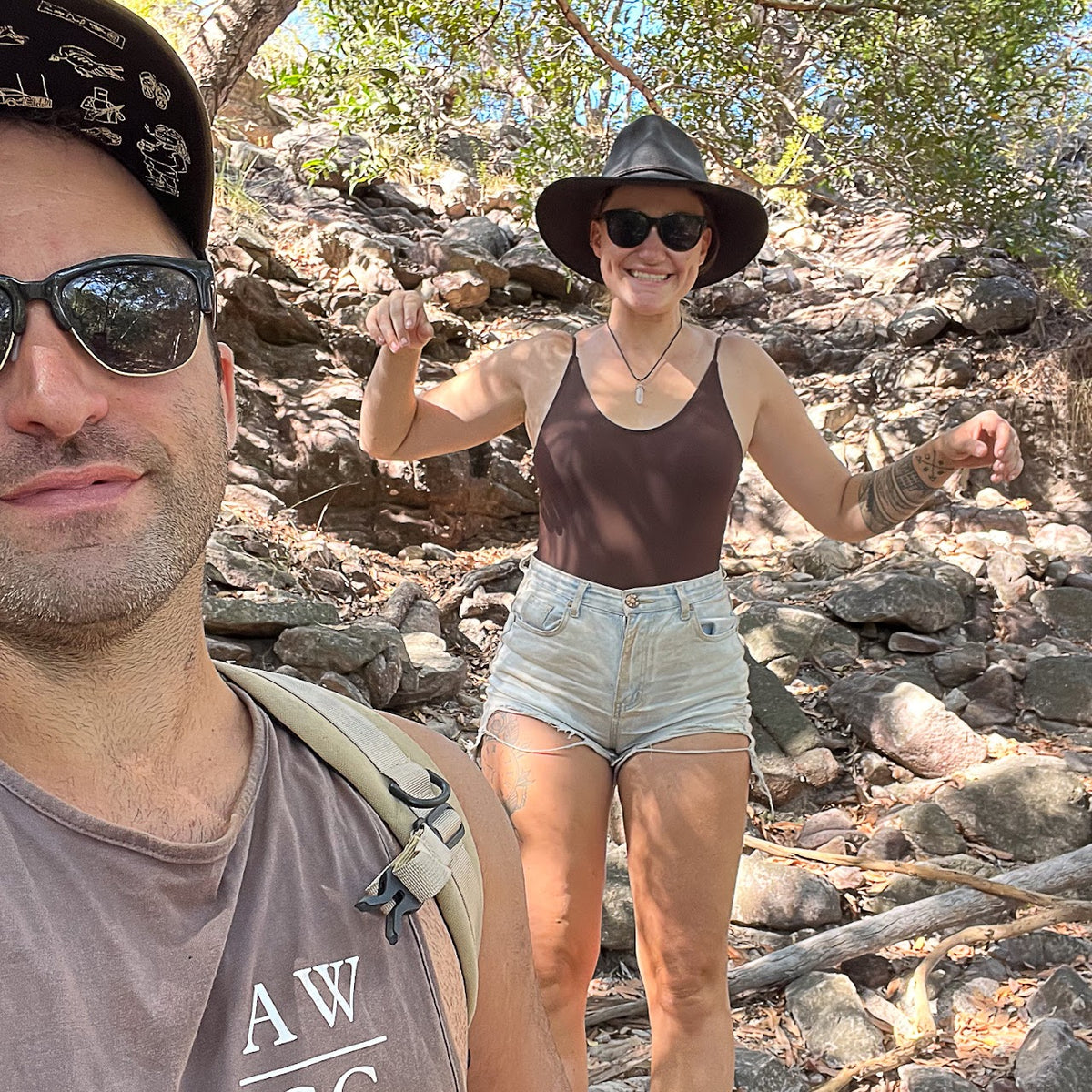 A man and a woman posing outdoors on a rocky, wooded trail. The woman is wearing a brown tank top, light denim shorts, a black wide-brim hat, and sunglasses, while the man in the foreground wears a brown t-shirt and a patterned black cap.