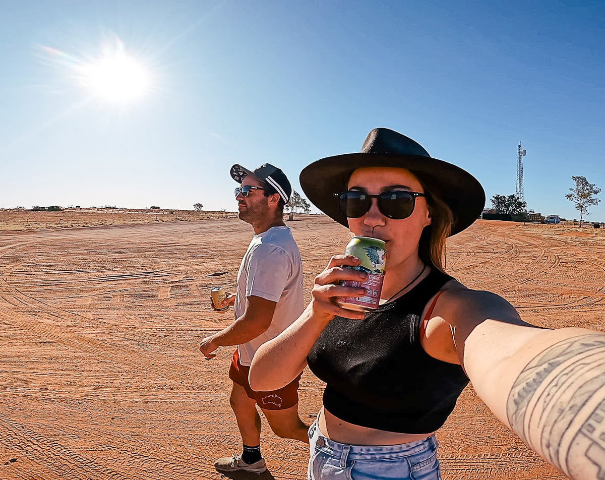 A woman in a black hat and sunglasses takes a selfie while holding a can of beer on a dusty red dirt road, with a man walking behind her under a bright sun and blue sky.