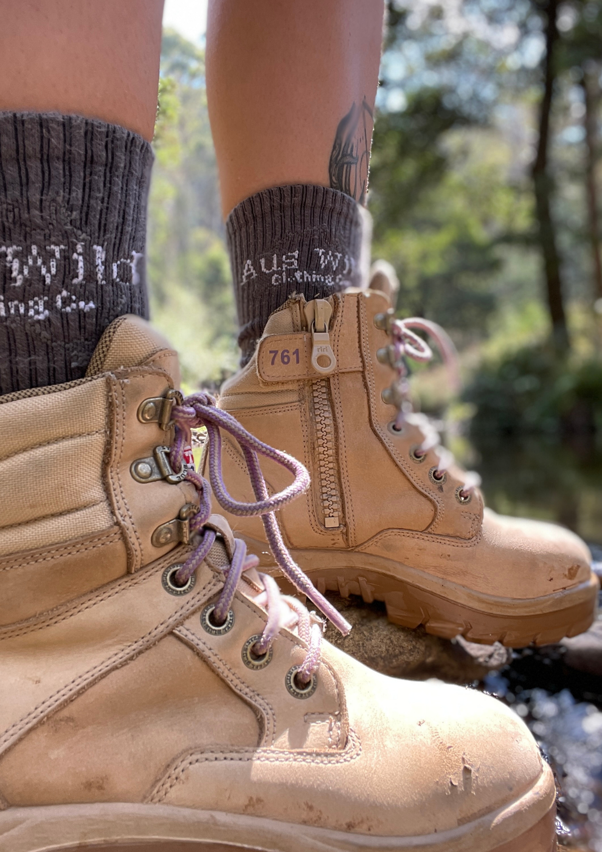 Close-up of a person wearing tan lace-up hiking boots with side zippers, dark gray socks, and standing outdoors near water with trees in the background.