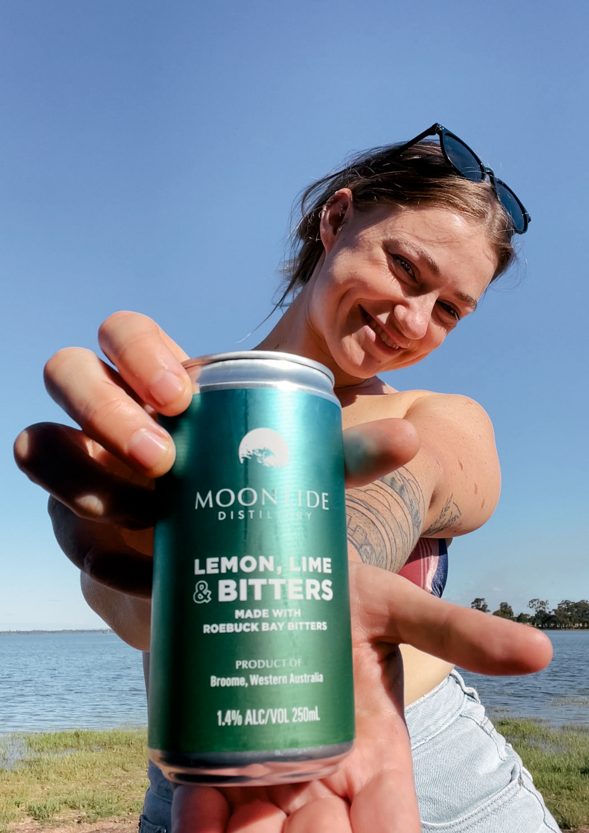 A woman outdoors by the water holds a green can of Moonside Distillery Lemon, Lime & Bitters toward the camera. The can is centered in the foreground, with the woman's face partially visible behind it against a blue sky and water background.